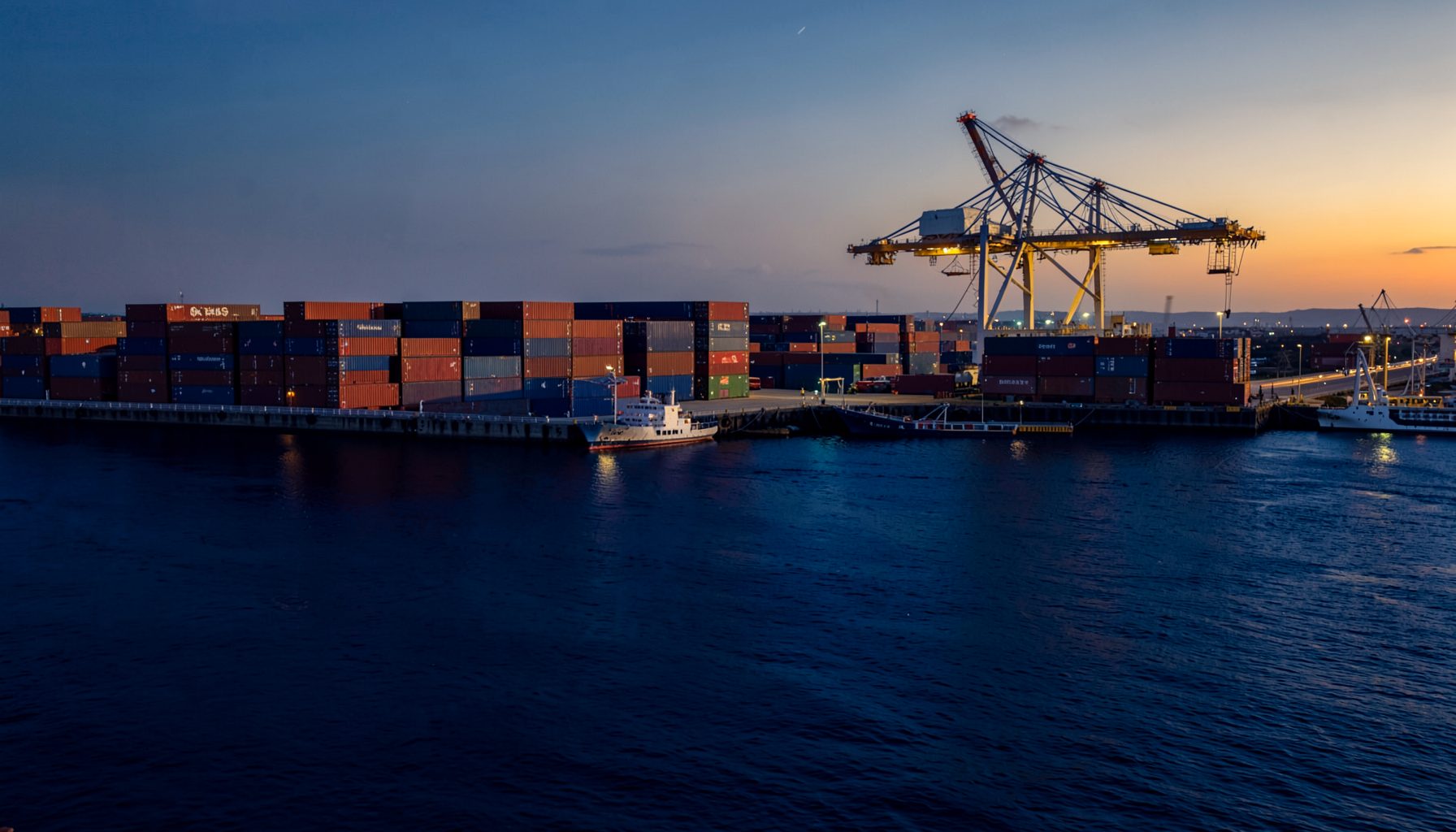 Wide harbor and cargo terminal scene in deep navy blue and warm golden light, with stacked shipping containers and a crane, evoking trade and logistics